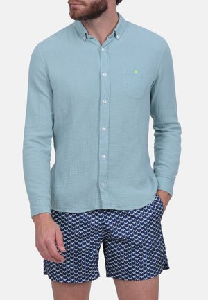 Chemise boutonnée bleu clair en tissu texturé, dotée d'une poche poitrine et de boutons blancs, associée à un short marine affichant un motif écaille de poisson.