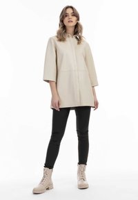 Beige, let shirt med krave, korte ærmer og knaplukning. Sammen med tætsiddende sorte bukser og beige snørebåndsstøvler.