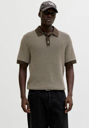 Młody mężczyzna w brązowej, wzorzystej dzianinowej koszulce polo, czarnych dżinsach i ciemnej czapce z białymi napisami, stojący na jednolitym tle.