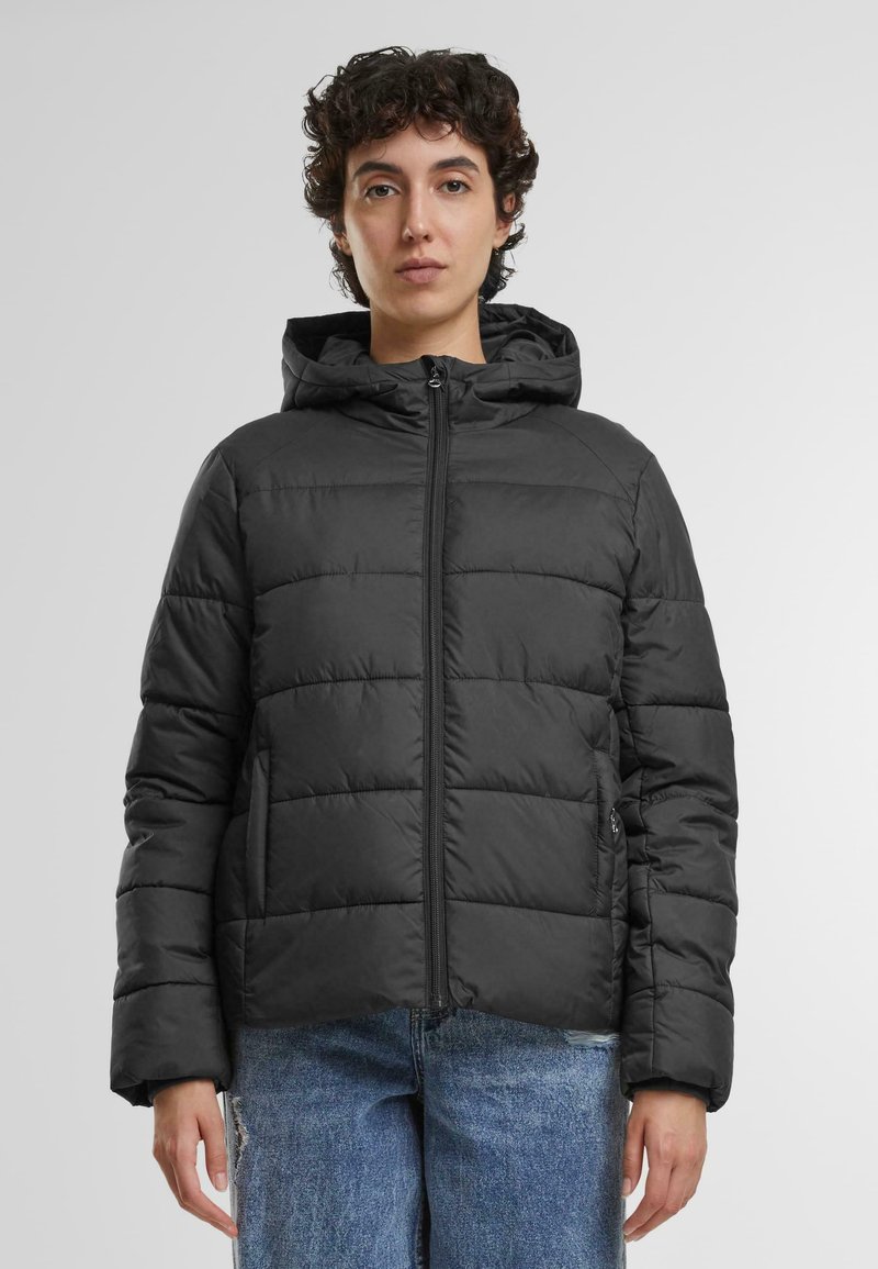 Puffjakke i ensfarget svart. Hettekonstruksjon med horisontal polstring, glidelås foran og sidelommer. Bært over uformelle blå jeans.