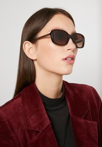 Gafas de sol en carey marrón con forma redondeada, lentes oscuros y varillas delgadas, llevadas por una persona con un blazer de pana burdeos.