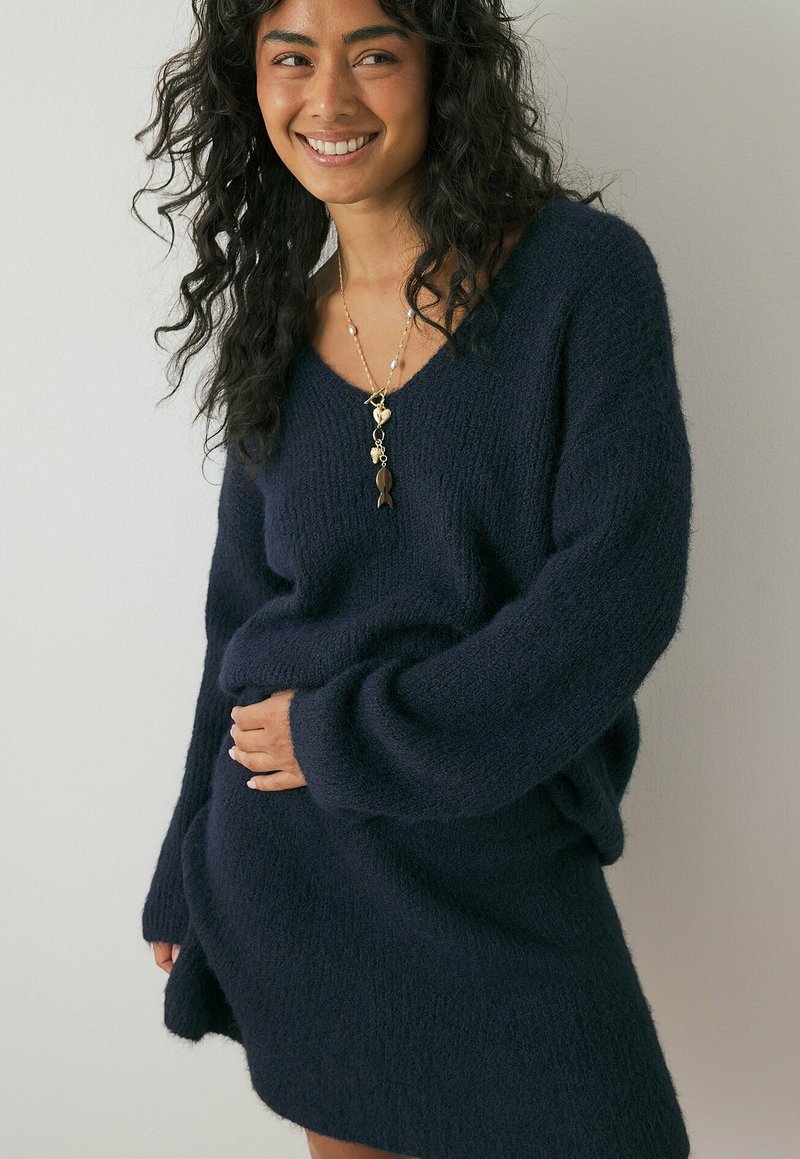 Navy knit sweater dress with a V-neck, oversized sleeves, and a soft, textured finish, worn with a layered gold necklace.