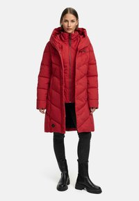 Rød lang pufferjakke med hætte, med et quiltet design, lynlås og trykknapper. Bæres over sorte jeans og sorte ankelstøvler.