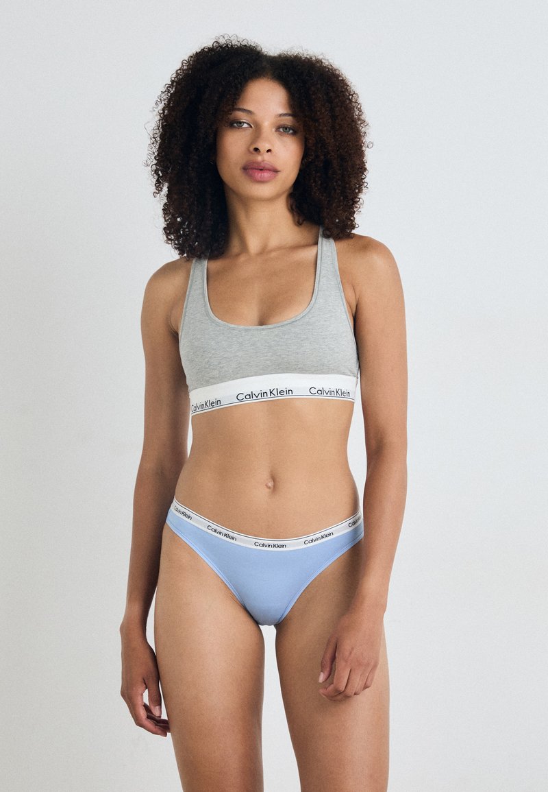 Pilkas sportiniai liemenėlė su Calvin Klein logotipo juostele; šviesiai mėlyni bikini apatiniai su atitinkama logotipo juostele. Audinys atrodo lygus ir elastingas.