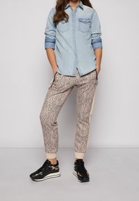 Ljus denimskjorta med två bröstfickor, kombinerad med mönstrade beige och bruna byxor. Svarta sneakers med metalliska detaljer.