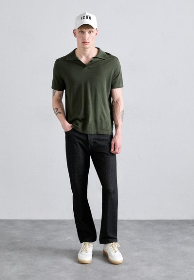 Grünes Poloshirt mit Kragen, kombiniert mit dunklen Jeans und weißen Sneakers. Das Modell trägt eine weiße Mütze mit dem Schriftzug "ICON".