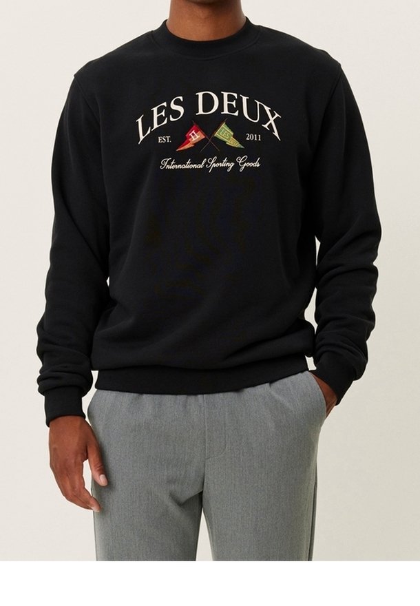 Homme portant un sweat-shirt noir avec le texte "LES DEUX" et deux petits drapeaux, associé à un pantalon gris clair.