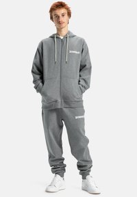 Grijze zip-up hoodie met crèmekleurige trekkoorden, gecombineerd met bijpassende joggingbroek. Beide items hebben het witte logo "DCSHOECO".