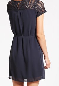 Námořnické modré šaty s krajkovými krátkými rukávy, skládacím sukní a knoflíkovým detailem na zadní straně. Vyrobeno z lehké tkaniny, která poskytuje volný střih.