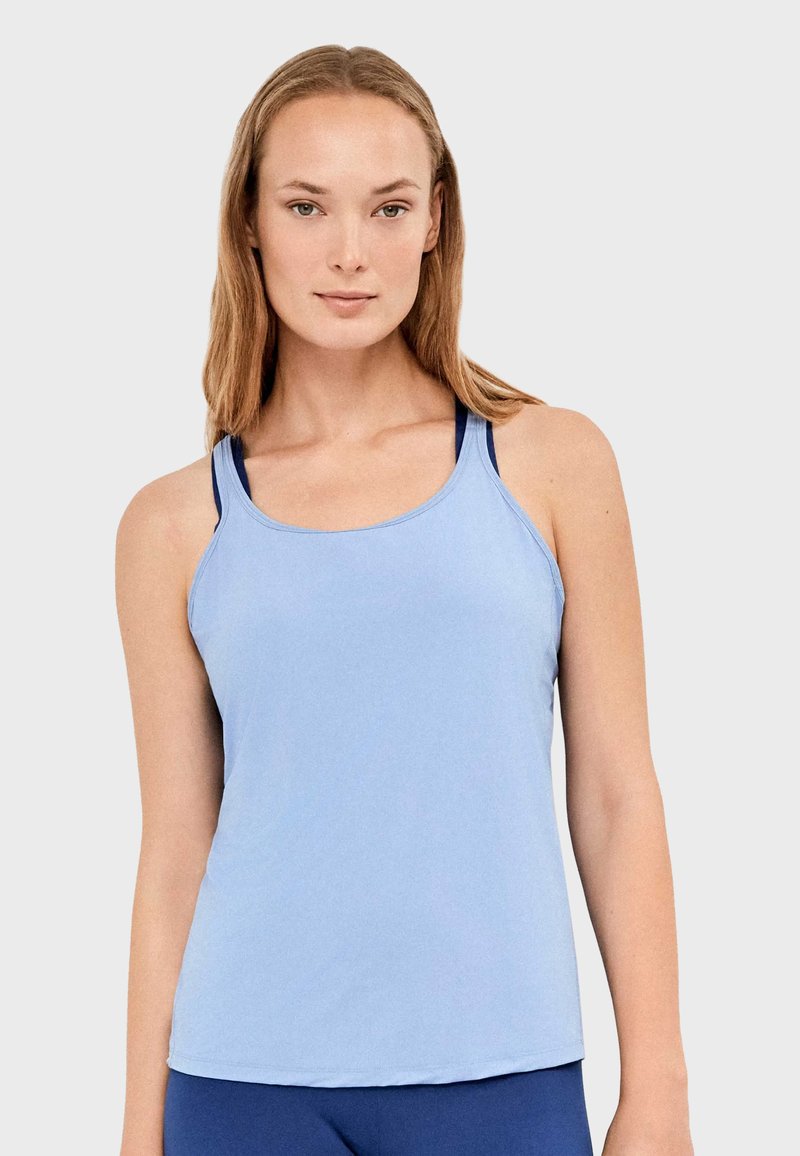 Jeune femme portant un débardeur sportif bleu clair sans manches par-dessus un soutien-gorge de sport foncé, posant devant un fond uni.