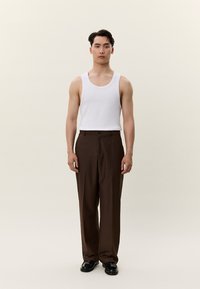 Mlad moški stoji, oblečen v belo majico brez rokavov, rjave krojene hlače in črne svečane čevlje, na preprostem svetlem ozadju.