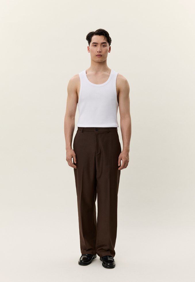 Mlad moški stoji, oblečen v belo majico brez rokavov, rjave krojene hlače in črne svečane čevlje, na preprostem svetlem ozadju.