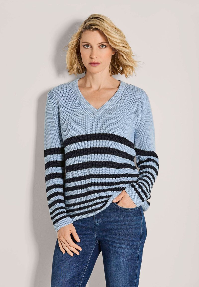 Frau mit blonden Haaren, die einen hellblauen Strickpullover mit schwarzen horizontalen Streifen und eine blaue Jeans trägt, steht vor einem schlichten hellen Hintergrund.
