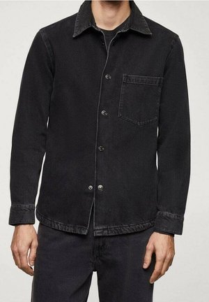 Mann trägt ein schwarzes Denim-Hemd mit Knopfleiste und Brusttasche, kombiniert mit schwarzen Hosen, steht vor einem schlichten hellen Hintergrund.