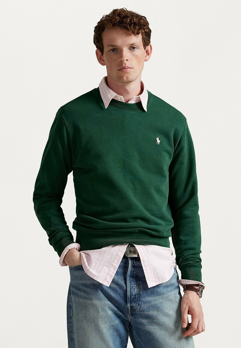 Maglione verde di cotone con scollo rotondo, indossato sopra una camicia a righe rosa chiaro con bottoni, abbinato a jeans di denim blu.