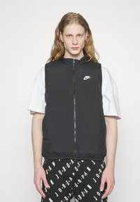 Crna prsluk bez rukava s visokim ovratnikom, patentnim zatvaračem i Nike logom. Nosi se preko bijele majice i crnih hlača s uzorkom ponovljenog loga.