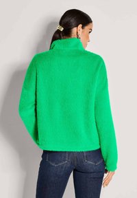 Frau mit dunklem Haar im Pferdeschwanz, die eine leuchtend grüne, flauschige Jacke und dunkelblaue Jeans trägt, mit dem Rücken zu einer schlichten hellen Hintergrund.