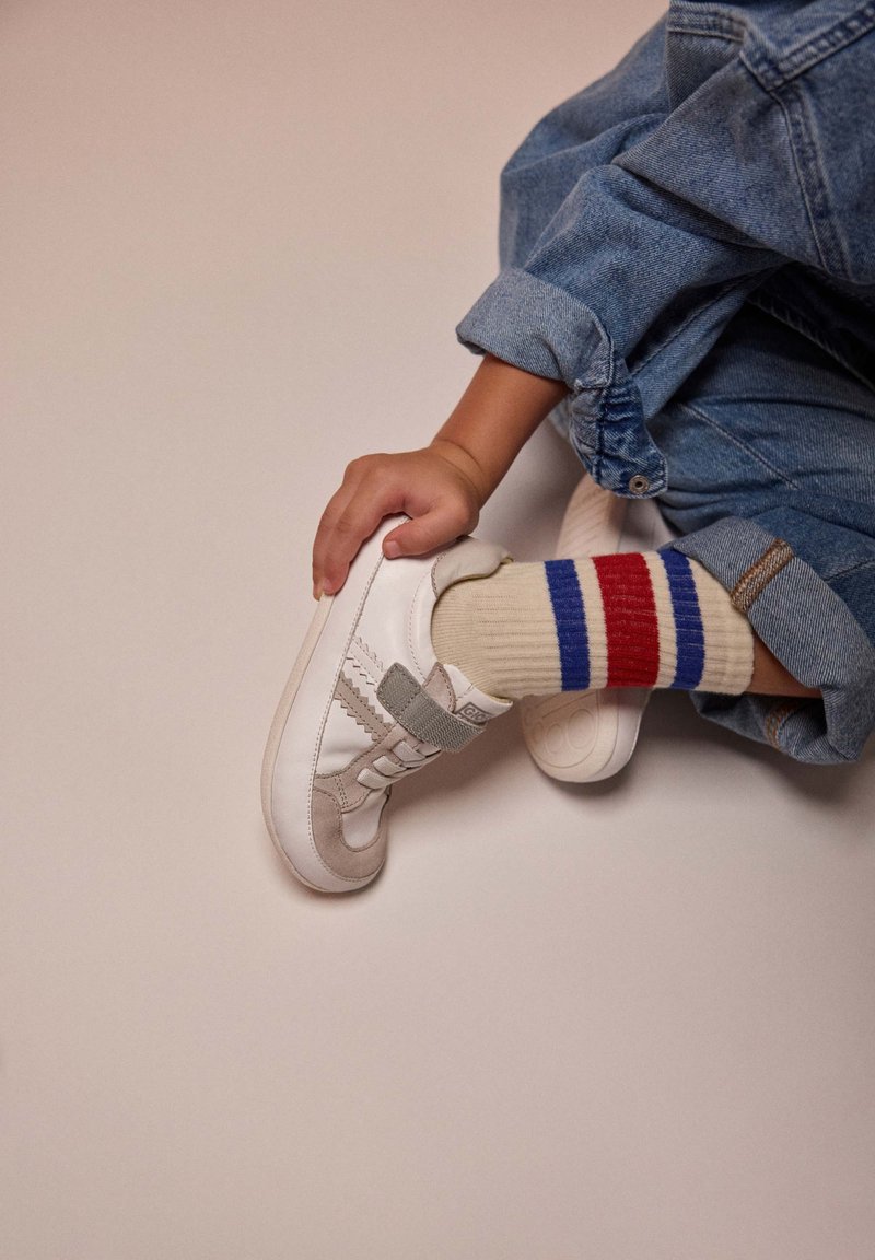 Baskets en cuir blanc avec des accents en daim gris, dotées d'une sangle velcro, accompagnées de chaussettes crème affichant des rayures rouges et bleues.