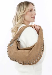 Bruine suede hobo tas met opvallende zilveren studs langs de naad, voorzien van een ritsluiting en een metalen bedel aan een ketting.