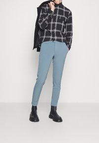 Černobílá kostičkovaná košile na knoflíky, světle modré slim-fit kalhoty a černé kotníkové boty. Hladká tkanina, přiléhavý střih, minimalistický design.