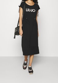 Czarna midi sukienka z krótkimi rękawami, z logo "LIUJO" w rzemieniu, z gumką w talii, zestawiona z czarnymi i w panterkę sneakersami.