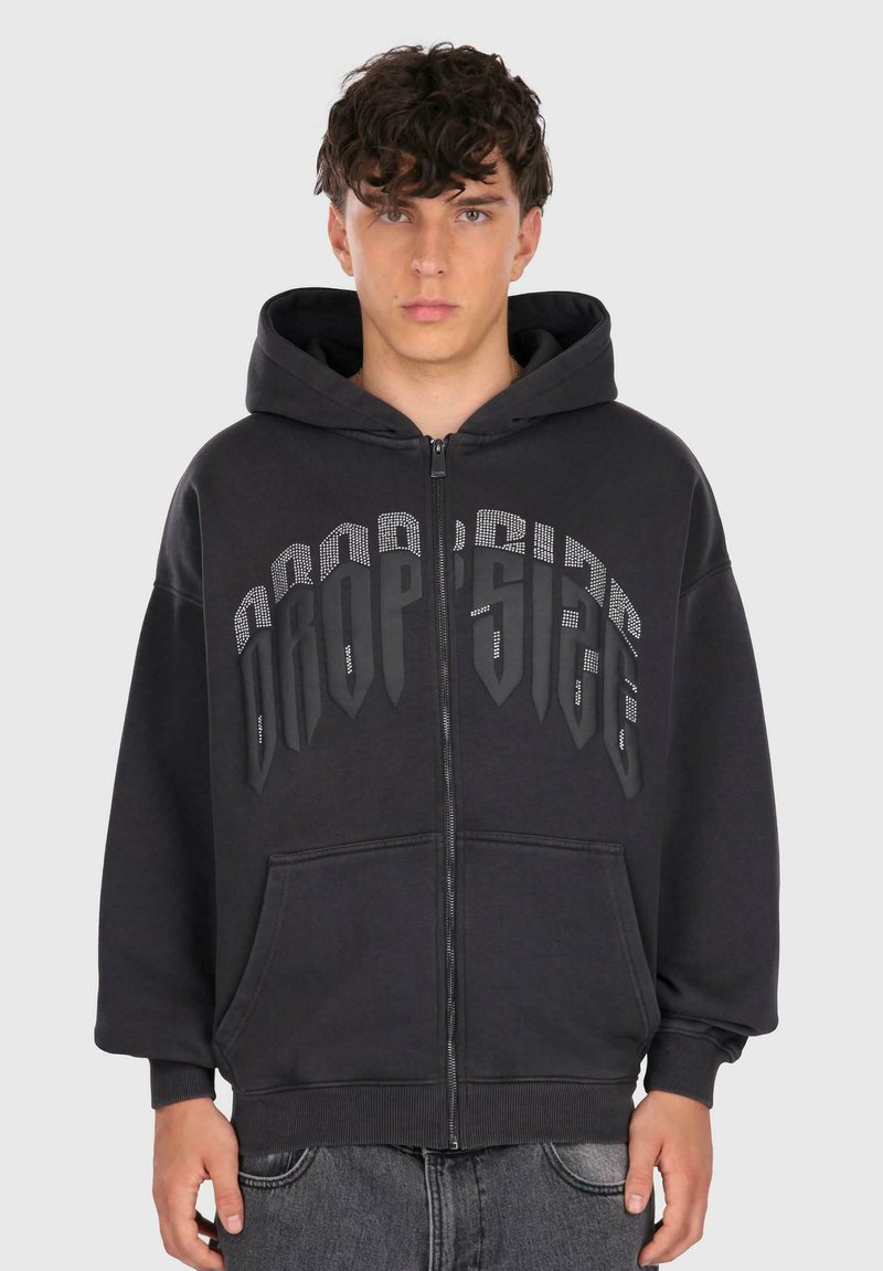 Ung mand iført en sort lynlås hættetrøje med stort "DROP SIDE" tekst i grå og hvide prikker over brystet, stående mod en ensfarvet baggrund.