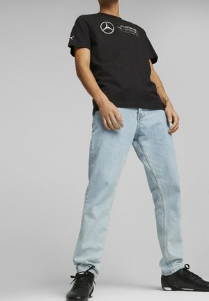 Persona vestida con una camiseta negra de Mercedes AMG Petronas, jeans de mezclilla azul claros y zapatillas deportivas negras, de pie contra un fondo gris plano.