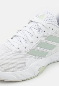 Scarpa da ginnastica bianca con tomaia in rete, lacci piatti e tre strisce laterali di un verde chiaro. La suola presenta dettagli testurizzati per una migliore aderenza.