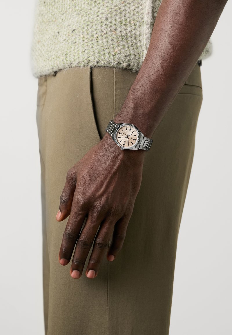 Montre en acier inoxydable avec un bracelet argenté et un cadran rose pâle, dotée d'index horaires noirs et d'une lunette texturée, portée sur une main.