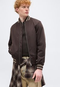 Νέος άνδρας με σγουρά μαλλιά που φοράει σκούρο καφέ bomber μπουφάν με φερμουάρ και μπεζ παντελόνι, με ένα πουκάμισο καρό δεμένο γύρω από τη μέση του.