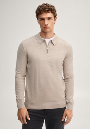 Giovane con capelli ricci che indossa un maglione beige con zip a maniche lunghe e pantaloni neri, in piedi contro uno sfondo semplice.
