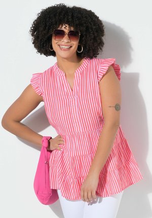 Smilende kvinde med krøllet hår iført firkantede solbriller, lyserødt og hvidt stribet ærmeløs top, hvide bukser og holder en lyserød taske.
