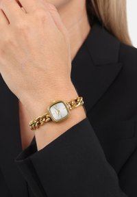 Goldener Armband-Uhr mit einem quadratischen weißen Zifferblatt, goldenen Stundenmarkierungen und einem massiven Gliederarmband, getragen an einem Handgelenk mit schwarzer Kleidung.