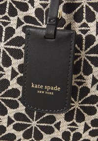 Fekete és krém színű, virágmintás szövet, egy téglalap alakú bőr címkével, melyen aranybetűkkel az áll; "kate spade NEW YORK".