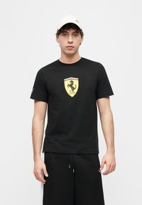 Νέος άνδρας που φοράει λευκό καπέλο και μαύρο T-shirt με κίτρινο λογότυπο ασπίδας με πηδηχτό άλογο, συνδυασμένο με μαύρα σορτς με κορδόνι και άσπρες πλευρικές ρίγες.