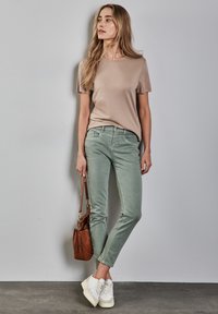 Camisa de manga corta beige, vaqueros slim-fit verde claro, zapatillas blancas con detalles grises y un bolso marrón. Conjunto simple y casual.