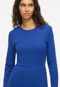 Modré šaty s dlouhým rukávem, žebrovanou texturou, kulatým výstřihem a rozšířenou sukní od pasu. Hladká látka bez viditelných vzorů.