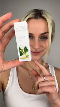 Femme aux cheveux blonds tenant et montrant une boîte de produit Kora Organics Noni Glow Face Oil avec une image botanique sur fond blanc.