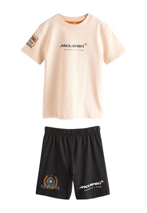 Jongere met lange dreadlocks die een beige McLaren Formula 1 Team T-shirt draagt, en de handen licht op schouderhoogte heft.