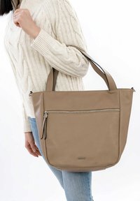 Beige leren tote bag met dubbele handvatten, een voorzak met rits en een gladde textuur. Geplaatst tegen een witte achtergrond en gedragen door een persoon.