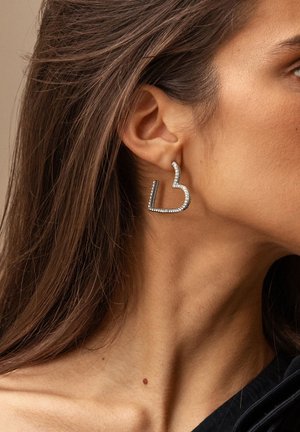 Pendientes de metal en tono plateado con forma de una "L" estilizada, adornados con cristales de imitación transparentes a lo largo de los bordes, llevados en la oreja de un modelo.