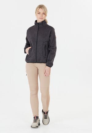 Kvinde stående iført en mørkegrå lynlåsjakke, beige bukser og beige vandresko mod en hvid baggrund.