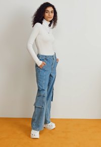 Hvid ribbet turtleneck-bluse, lyseblå cargo-bukser med lommer, afslappet pasform, og hvide slip-on sko på orange baggrund.