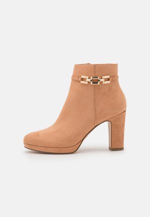 Tan suède enkellaarsje met blokhak en goudkleurige decoratieve gesp aan de zijkant tegen een effen achtergrond.