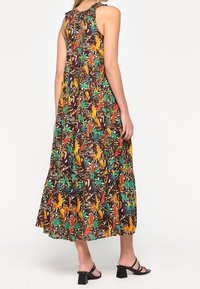 Maxi šaty bez rukávů s jasným tropickým vzorem ptáků a leopardími skvrnami, nosící černé sandály na blokovém podpatku se řemínky.