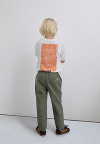 Niño con cabello rubio corto se encuentra frente a una pared, vistiendo una camiseta blanca con un gráfico de cancha de baloncesto en naranja y pantalones verde oliva.