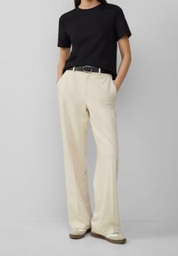 Černé tričko s kulatým výstřihem spojené s krémovými kalhotami s širokými nohavicemi, zdobené páskem se stříbrnou sponou a béžovými botami se šedými akcenty.