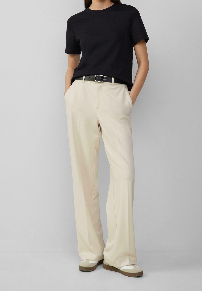 Černé tričko s kulatým výstřihem spojené s krémovými kalhotami s širokými nohavicemi, zdobené páskem se stříbrnou sponou a béžovými botami se šedými akcenty.