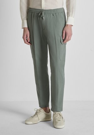 Persona che indossa pantaloni cargo verde chiaro con tasche frontali e sneakers beige, in piedi contro uno sfondo grigio uniforme.