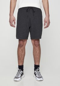 Shorts gris texturés avec une taille élastique et un cordon de serrage. Portés avec des baskets grises et des chaussettes foncées, debout devant un fond uni.